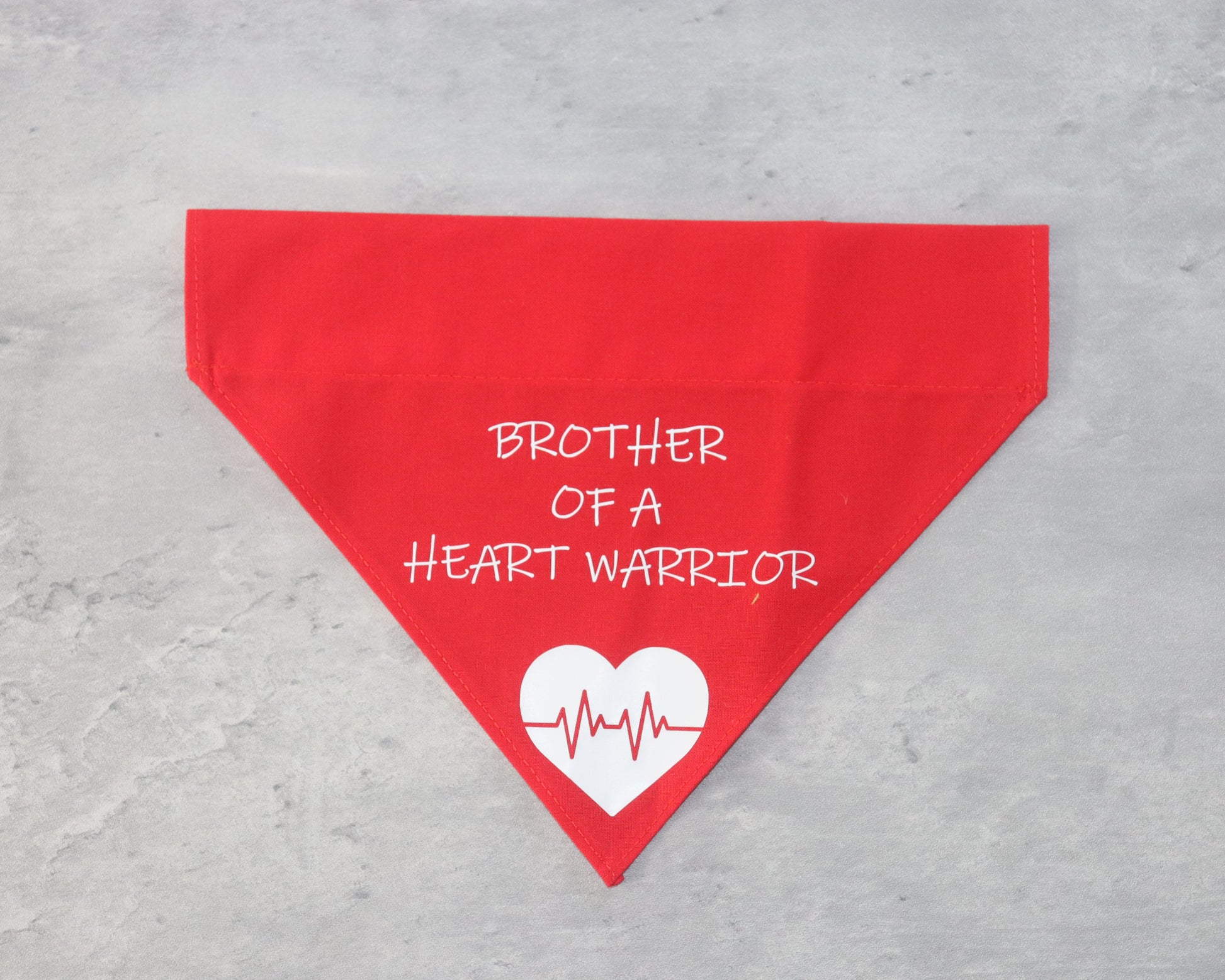 A red triangular bandana with the text 'BROTHER OF A HEART WARRIOR' printed in white. Below the text, there is a white heart graphic with a red heartbeat line running through it. The bandana is laid flat on a gray surface.