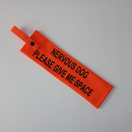 A fluorescent orange fabric lead sleeve with a black button snap. The sleeve features bold black text reading 'NERVOUS DOG PLEASE GIVE ME SPACE.' Designed to help dog owners signal that their pet needs space, this high-visibility accessory prevents unwanted interactions and helps ensure a stress-free environment for nervous dogs.