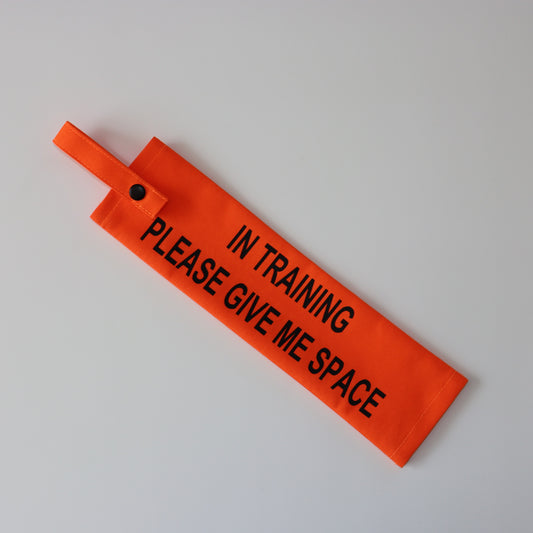 A fluorescent orange fabric lead sleeve with a black button snap. The sleeve displays bold black text reading 'IN TRAINING PLEASE GIVE ME SPACE.' Designed for dog handlers managing pups in training, its bright color and clear text ensure visibility. Ideal for service dogs, working dogs, or pets in training, this sleeve helps communicate to passersby that the dog should not be distracted or engaged with."
