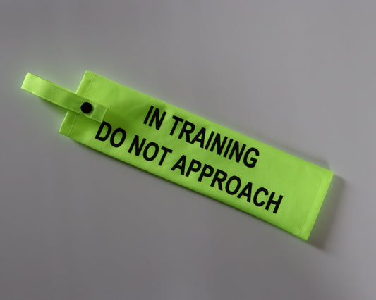 A fluorescent yellow fabric lead sleeve with a black button snap. The sleeve features bold black text that reads 'IN TRAINING DO NOT APPROACH.' Designed for dog walkers handling pups in training, the bright colour and clear typography ensure the message is highly visible, helping prevent unwanted interactions while allowing the dog to focus on their development.