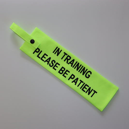 A fluorescent yellow fabric lead sleeve with a black button snap. The sleeve features bold black text that reads 'IN TRAINING PLEASE BE PATIENT.' Designed specifically for dog walkers handling pups in training, its bright colour and clear typography ensure the message is highly visible, helping to encourage patience and understanding from passersby while allowing the dog to focus on their development.
