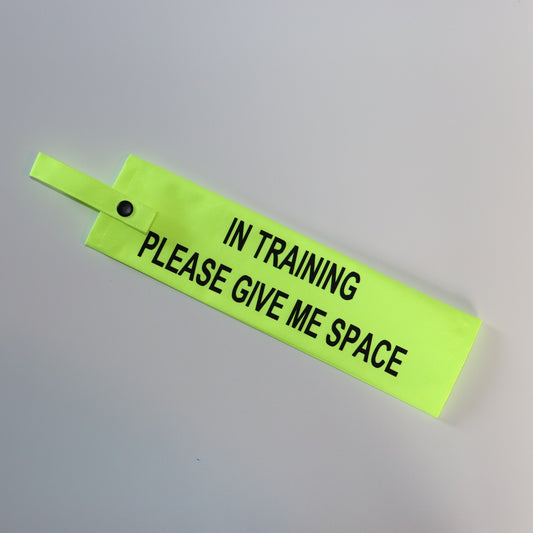 A fluorescent yellow fabric lead sleeve with a black button snap. The sleeve displays bold black text reading 'IN TRAINING PLEASE GIVE ME SPACE.' Designed for dog handlers managing pups in training, its bright color and clear text ensure visibility. Ideal for service dogs, working dogs, or pets in training, this sleeve helps communicate to passersby that the dog should not be distracted or engaged with.