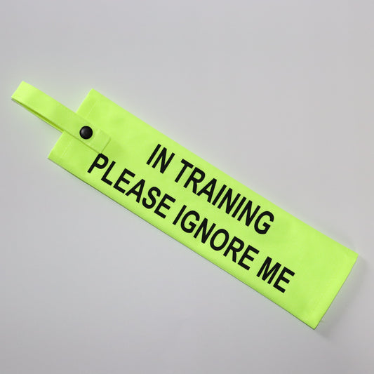 A fluorescent yellow fabric lead sleeve with a black button snap. The sleeve displays bold black text reading 'IN TRAINING PLEASE IGNORE ME.' Designed for dog handlers managing pups in training, its bright color and clear, authoritative text ensure visibility. Ideal for service dogs, working dogs, or pets in training, this sleeve helps communicate to passersby that the dog should not be distracted or engaged with."