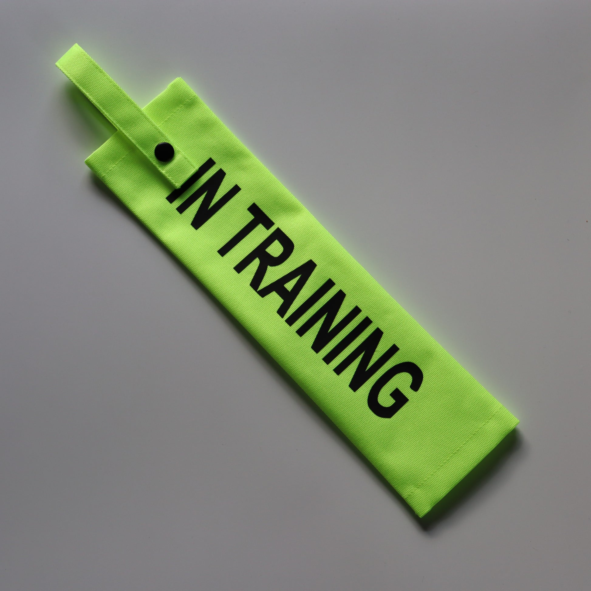 A fluorescent yellow fabric lead sleeve with a black button snap. The sleeve displays bold black text reading 'IN TRAINING.' Designed for dog walkers handling pups in training, its high-visibility colour and clear typography ensure the message is noticeable, helping to inform passersby that the dog is in the process of learning and development.
