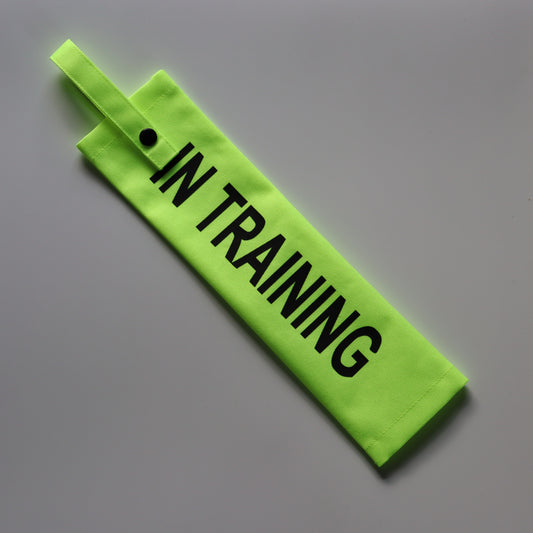 A fluorescent yellow fabric lead sleeve with a black button snap. The sleeve displays bold black text reading 'IN TRAINING.' Designed for dog walkers handling pups in training, its high-visibility colour and clear typography ensure the message is noticeable, helping to inform passersby that the dog is in the process of learning and development.