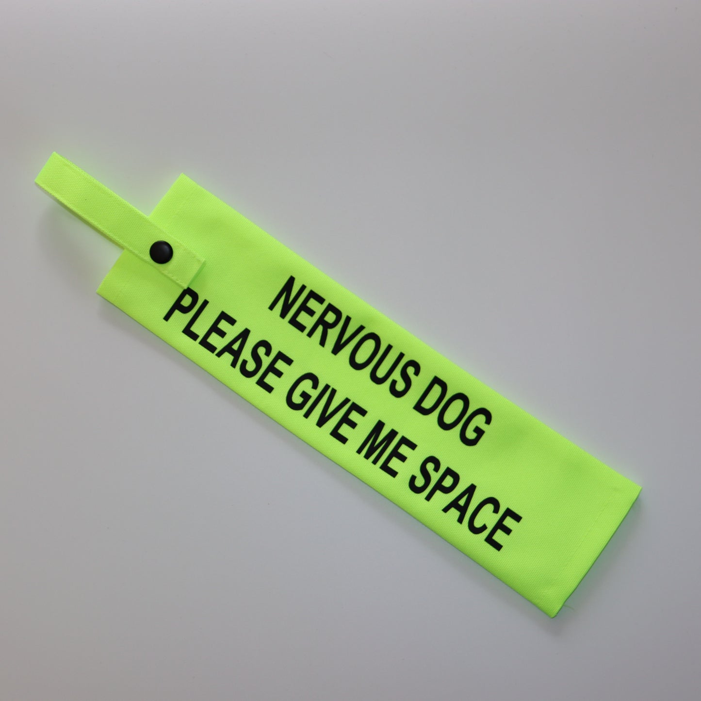 A fluorescent yellow fabric lead sleeve with a black button snap. The sleeve features bold black text reading 'NERVOUS DOG PLEASE GIVE ME SPACE.' Designed to help dog owners signal that their pet needs space, this high-visibility accessory prevents unwanted interactions and helps ensure a stress-free environment for nervous dogs.