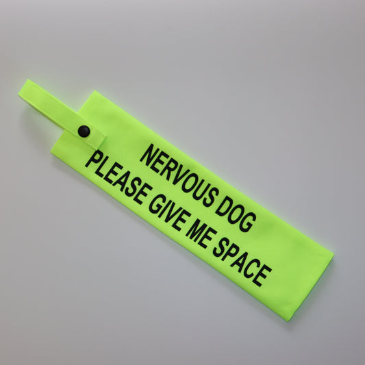A fluorescent yellow fabric lead sleeve with a black button snap. The sleeve features bold black text reading 'NERVOUS DOG PLEASE GIVE ME SPACE.' Designed to help dog owners signal that their pet needs space, this high-visibility accessory prevents unwanted interactions and helps ensure a stress-free environment for nervous dogs.