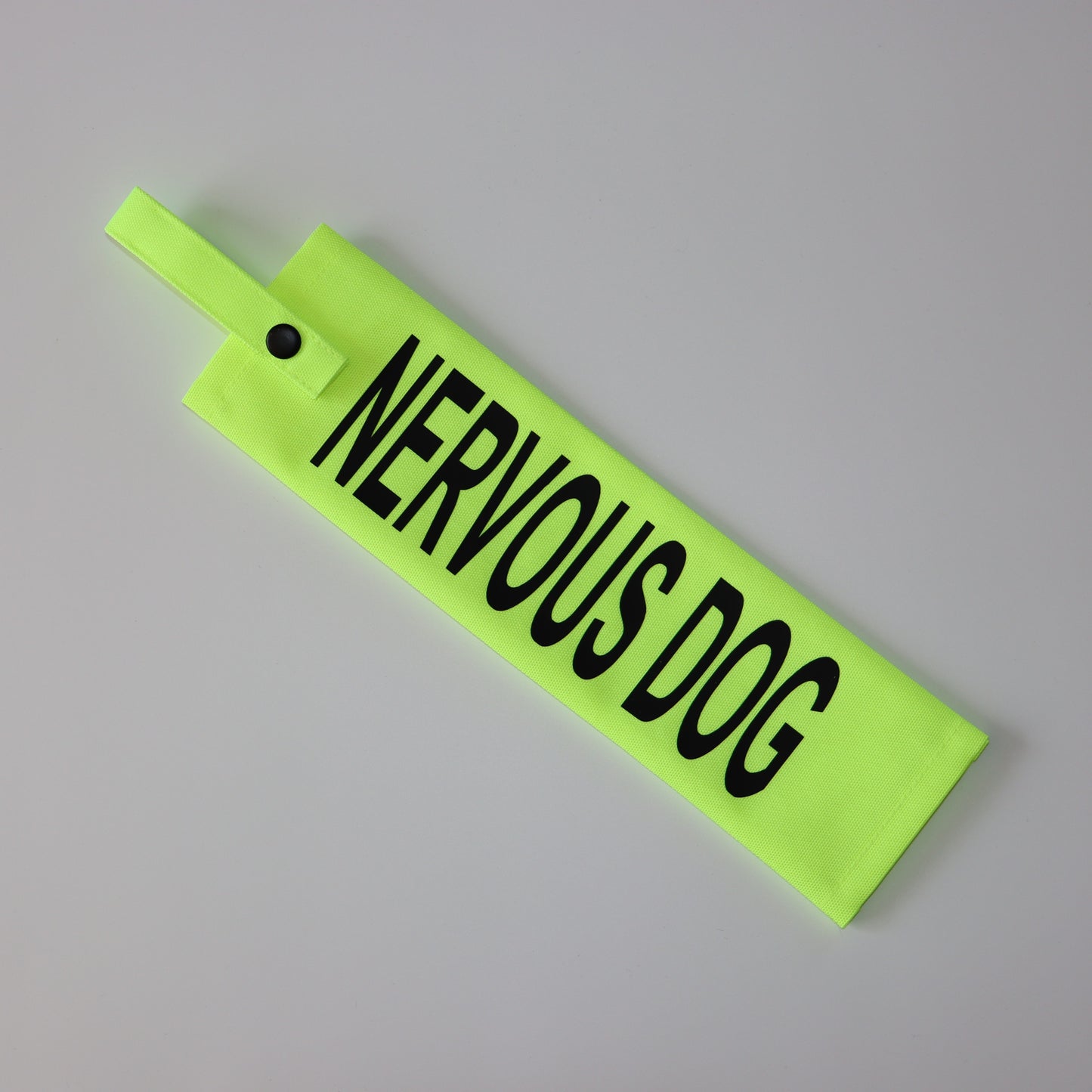 A fluorescent yellow fabric lead sleeve with a black button snap. The sleeve displays bold black text reading 'NERVOUS DOG.' Designed to help dog owners communicate their pet’s anxiety, this sleeve ensures clear visibility with its bright color and bold lettering. Ideal for dogs that require space, the high-visibility design helps prevent stressful interactions and unwanted approaches.