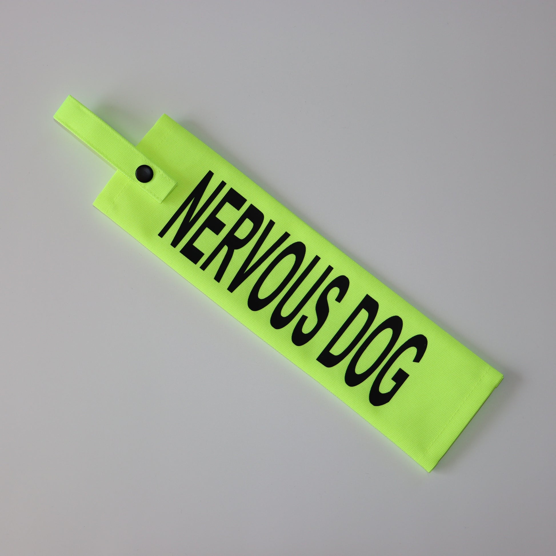 A fluorescent yellow fabric lead sleeve with a black button snap. The sleeve displays bold black text reading 'NERVOUS DOG.' Designed to help dog owners communicate their pet’s anxiety, this sleeve ensures clear visibility with its bright color and bold lettering. Ideal for dogs that require space, the high-visibility design helps prevent stressful interactions and unwanted approaches.