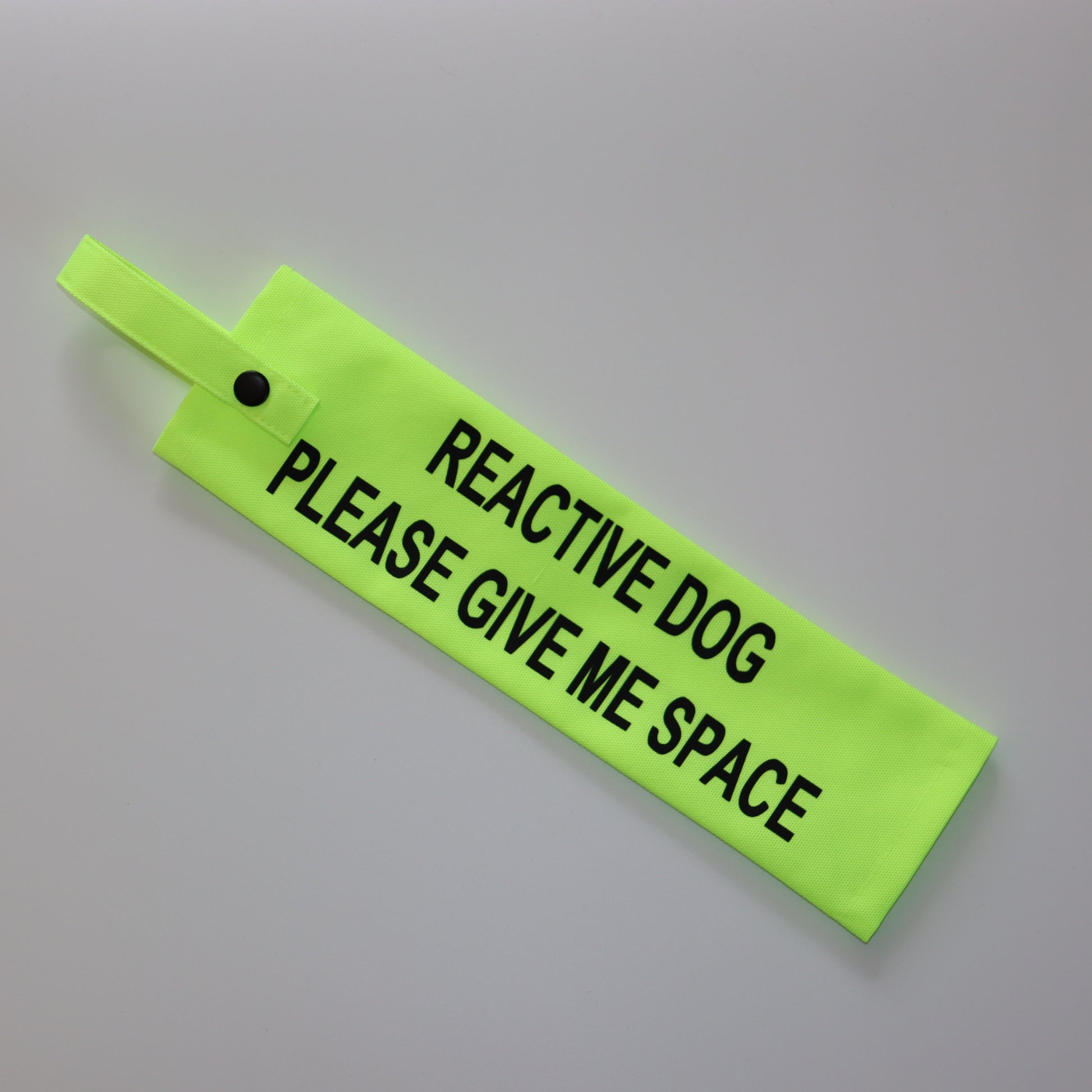 A fluorescent yellow fabric lead sleeve with a black button snap. The sleeve features bold black text reading 'REACTIVE DOG PLEASE GIVE ME SPACE.' Designed to help dog owners signal that their pet needs space, this high-visibility accessory prevents unwanted interactions and helps ensure a stress-free environment for dogs prone to reactive behavior.