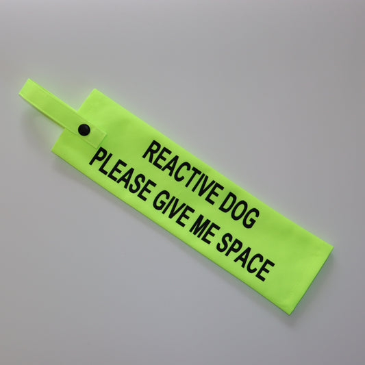 A fluorescent yellow fabric lead sleeve with a black button snap. The sleeve features bold black text reading 'REACTIVE DOG PLEASE GIVE ME SPACE.' Designed to help dog owners signal that their pet needs space, this high-visibility accessory prevents unwanted interactions and helps ensure a stress-free environment for dogs prone to reactive behavior.